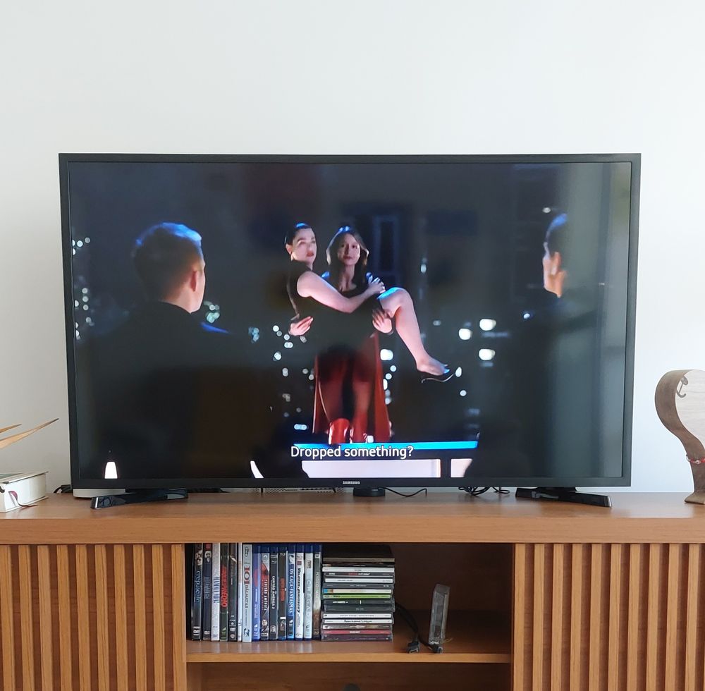 Foto de uma televisao exibindo um episodio de Supergirl em que ela carrega Lena Luthor nos braços como se fosse uma noiva.