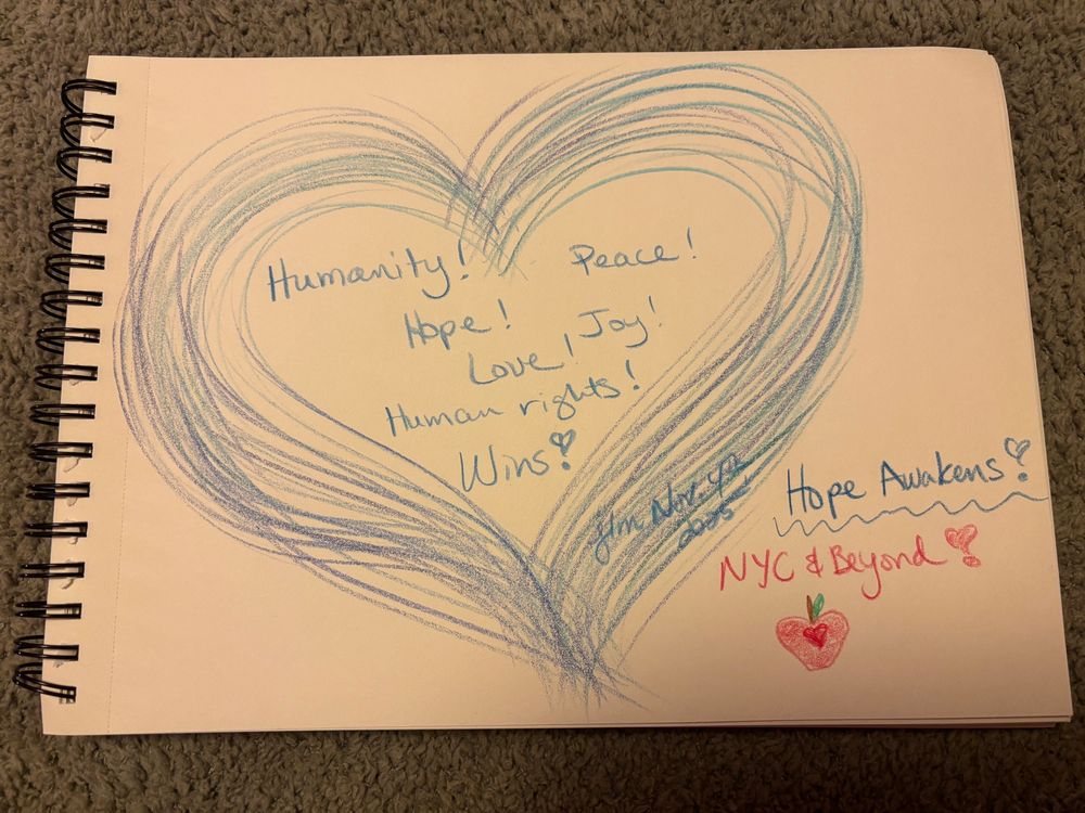 My drawing with mostly various shades of blue colored pencils. A multi colored blue heart with the words Humanity! Peace! Hope! Joy! Love! Human Rights! Wins! Hope Awakens! And in red colored pencil, NYC & Beyond! And a tiny red drawing of a heart shaped apple. 
