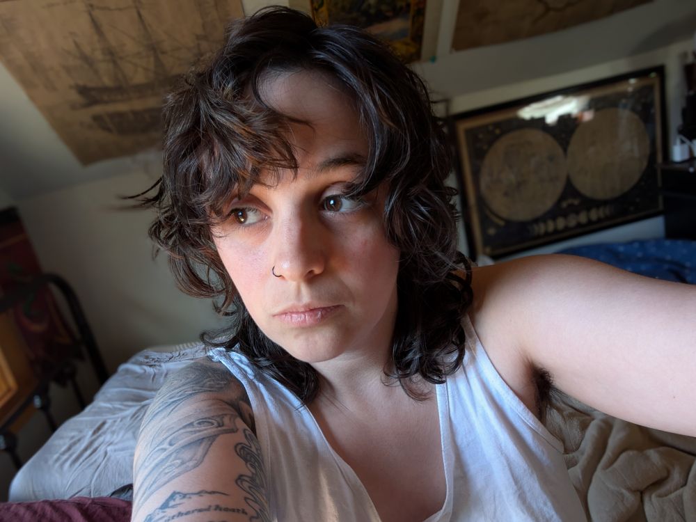 Selfie of a nonbinary white person with brown-black hair cut about shoulder length, brown eyes looking off to one side, and wearing a white tank top. There are visible tattoos on their right shoulder. The hair style is akin to 70s George Harrison
