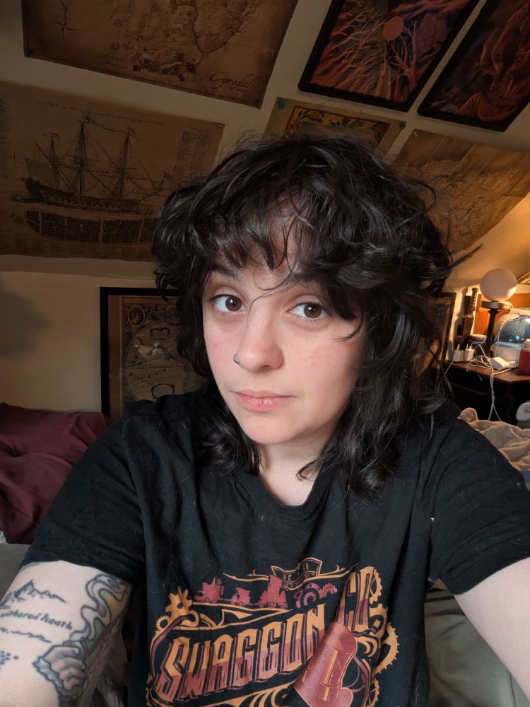 Selfie of me, a white non-binary person with brown eyes and black-brown hair. I have a small hoop in one nostril and visible tattoos on one arm, and I am wearing a good times with scar shirt. My face is trying to look chill but I look visibly tired