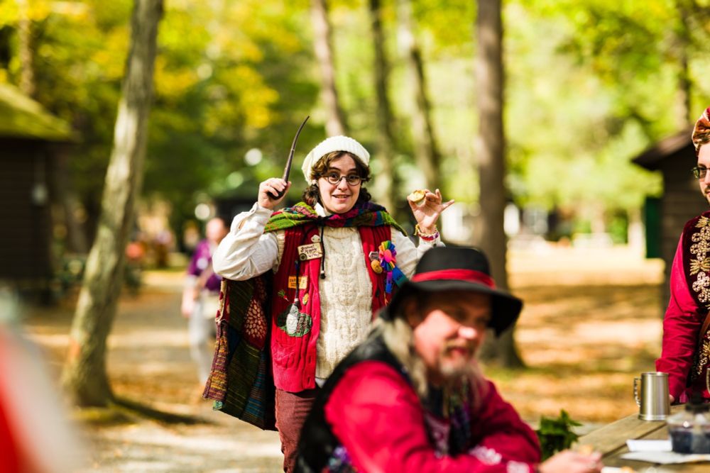 Picture from Shirefolk larp. JeraWizard is wearing warm-looking hobbit clothing, including a patchwork cape, white knit hat, and a red vest with many patches on it. They are holding a long pipe in one hand, and a muffin in the other, and appear to be mid-rant.