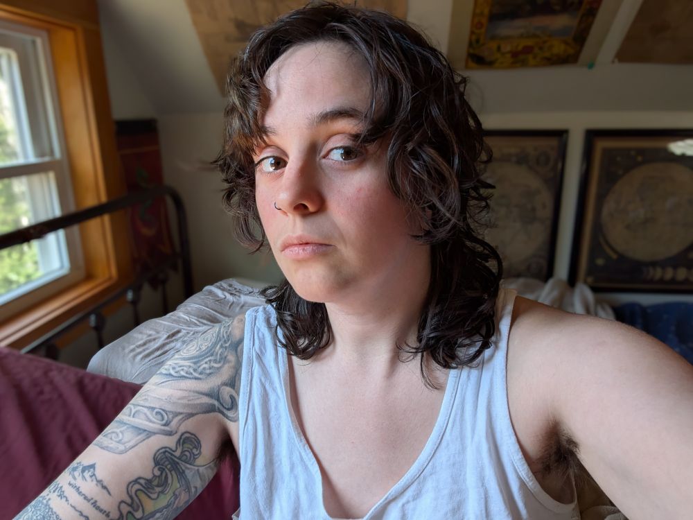 Selfie of a nonbinary white person with brown-black hair cut about shoulder length, brown eyes looking into the camera, and wearing a white tank top. There are visible tattoos on their right shoulder. The hair style is akin to 70s George Harrison