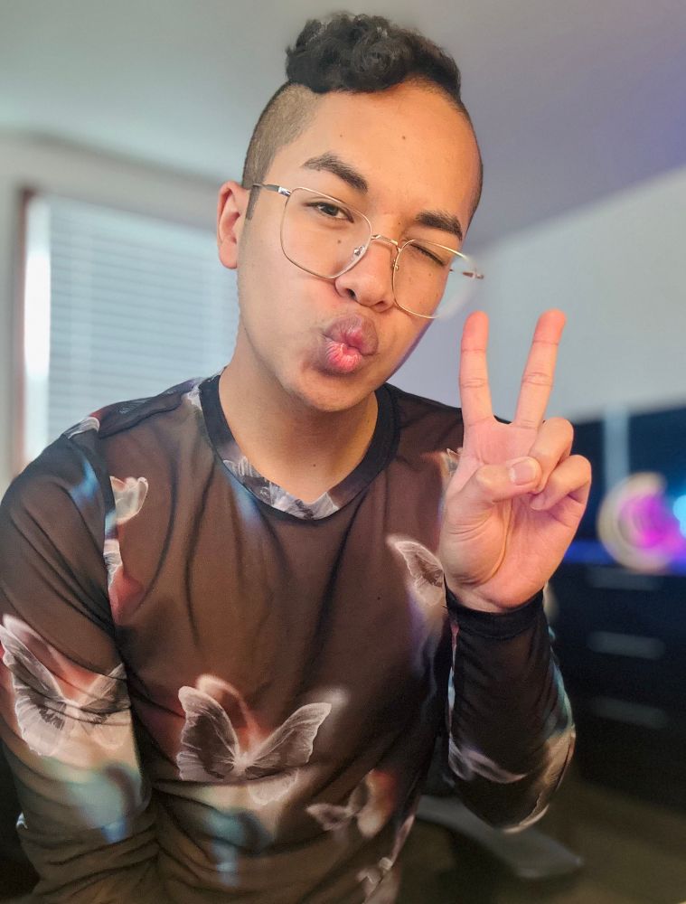 Sass posing with duck lips, a wink on the right eye, while holding up a peace sign. He’s posing towards the camera while sitting and wearing a black mesh shirt with white/colorful butterfly detailing