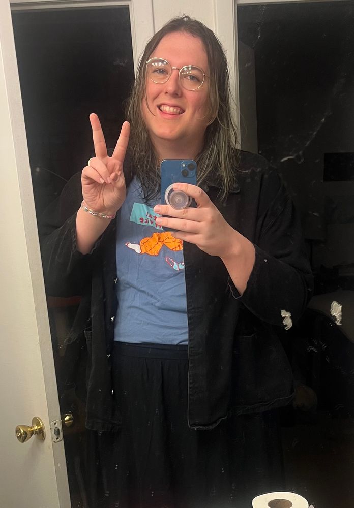 a bathroom selfie of a white trans woman (me!) with shoulder length hair and glasses in a black skirt with a blue ‘the postal service’ band t-shirt