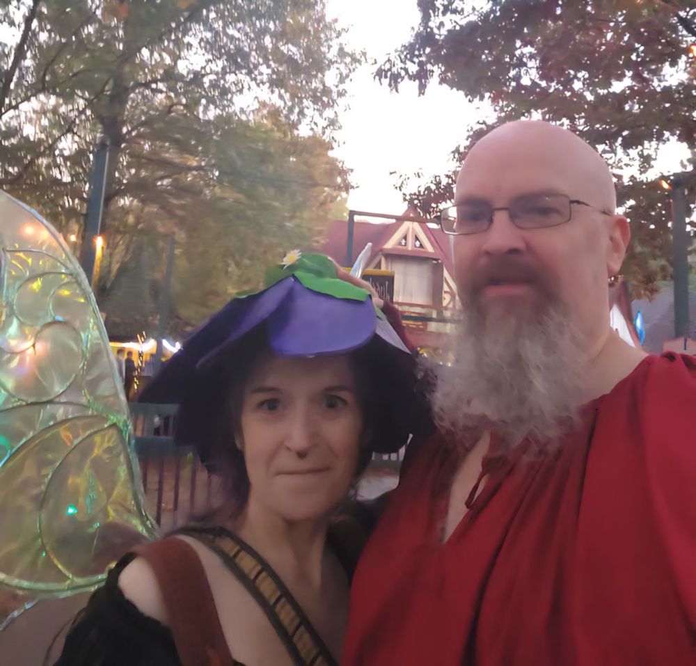 At the Georgia Renaissance Faire's Fall Fling with myself on the left & SO on the right. I'm dressed in a purple flower hat, green iridescent faire wings and an of the shoulder green top. SO has a long white goatee and a puffy, red,  pirate shirt. 