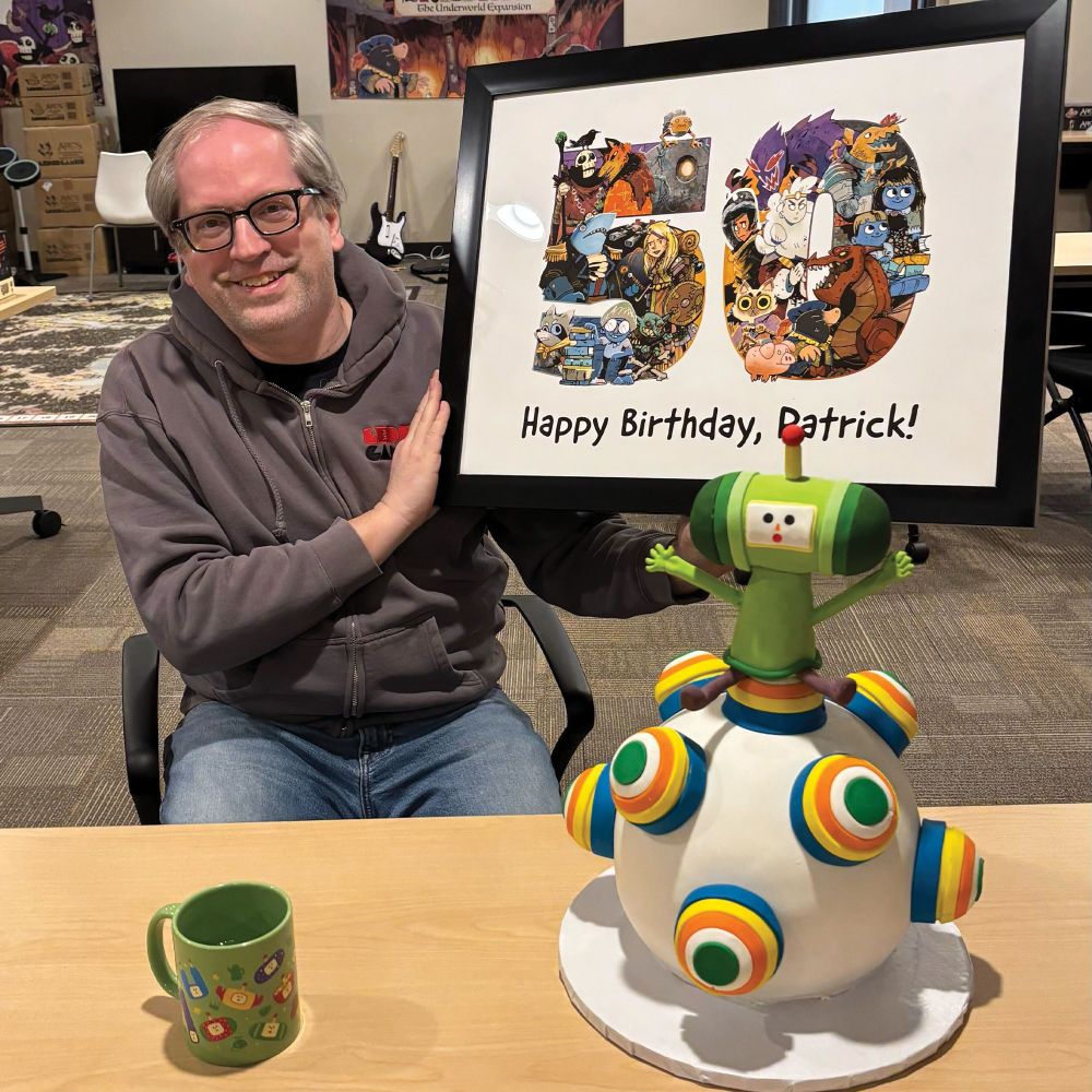 Patrick Leder holds up a print that says Happy Birthday Patrick and "50" made up of characters from Leder Games. On the table there is a figural cake featuring a Katamari ball and the Prince. 
