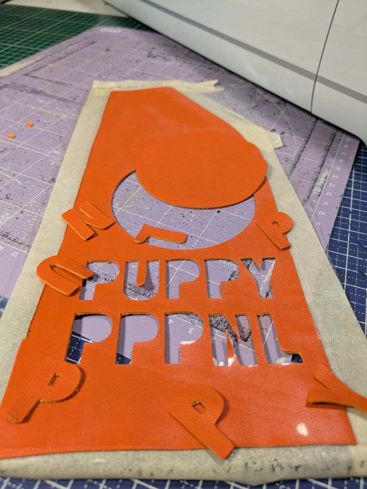 A piece of orange leather with tape around it and the following shapes cut out of it and laying on top of it, a circle and the letters P U P P Y N L