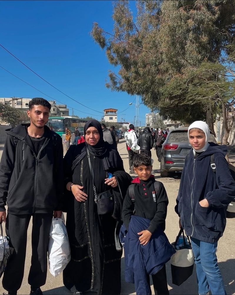 A picture of Maya Mohareb and her family in Gaza. They are standing in a street on a sunny day, wearing mostly black and blue. From left to right: her older brother, her mother, her younger brother and Maya.  They are facing the camera with small hopeful smiles.  