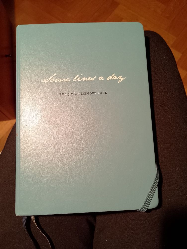 Poor quality photo of a blue notebook, embossed with the writing
Some lines a day (write, cursive) 
The 5 year notebook (black, small caps) 
One can glimpse an elastic band to keep the notebook closed, and two cloth bookmarks, one dark blue and one grey. 
