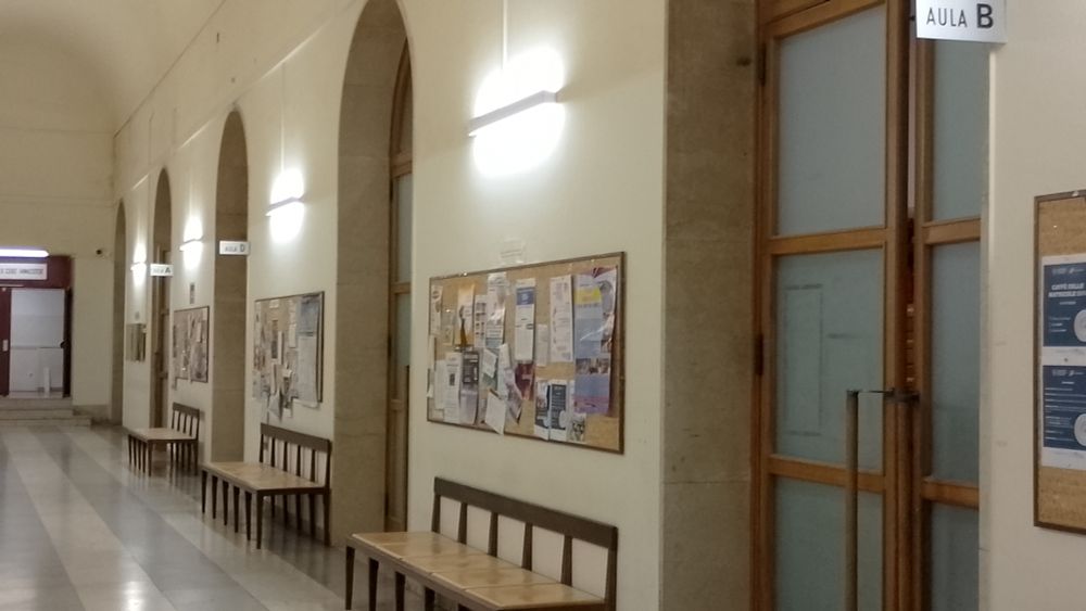 A corridor with wooden benches and artificial light. On the side one can see four large wooden doors labelled in order Aula B, No Label, Aula D, Aula A.