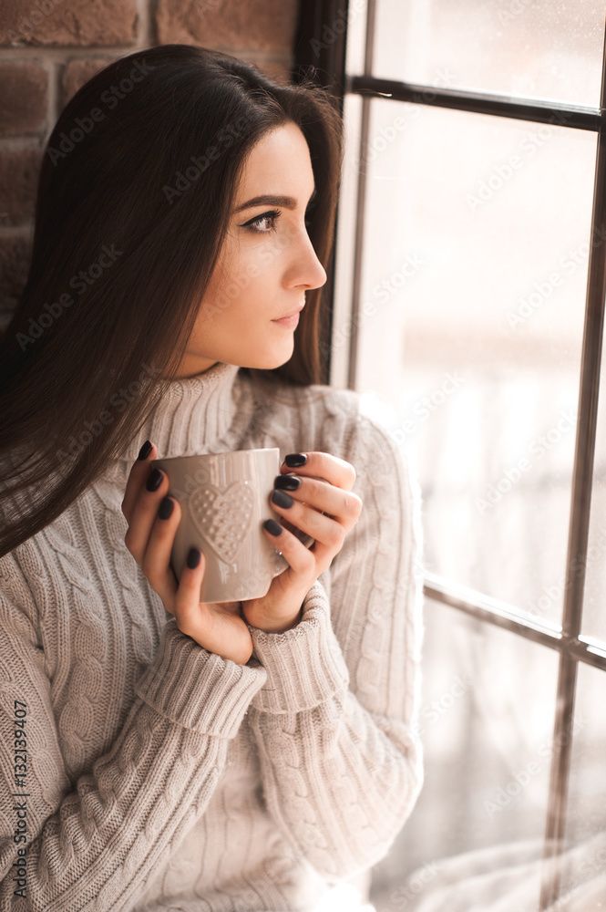 A cheugy white woman in a chunky sweater holding a hot mug of coffee next to a window