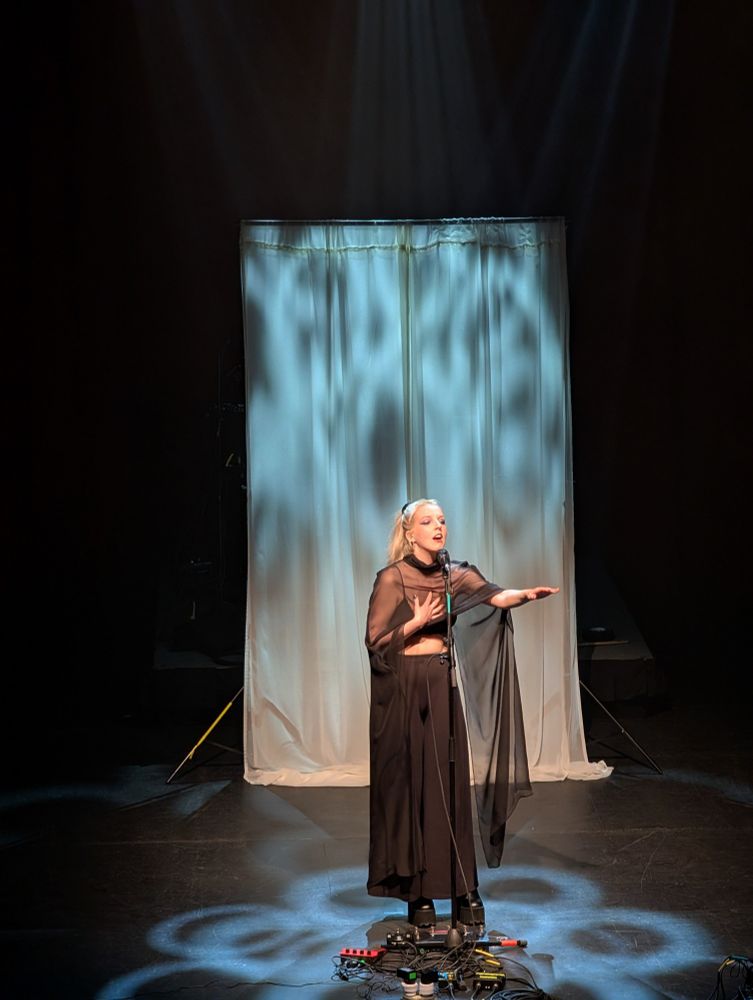 The singer Sylvaine, alone on stage, singing in front of a backdrop, surrounded by bluish-white stage lights.