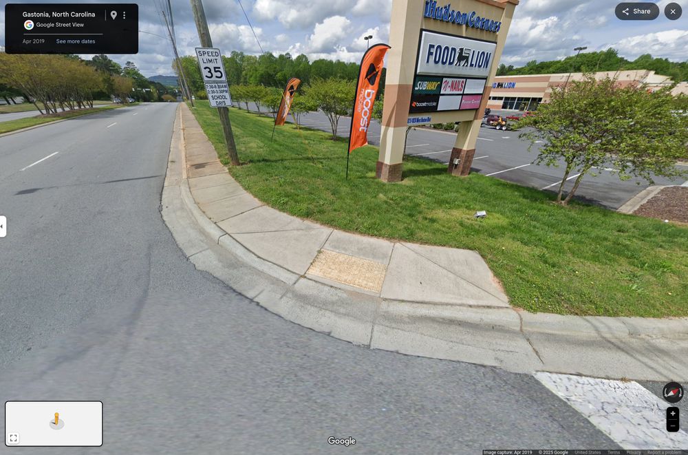 Image of the corner on W Hudson where the grocery store ("Hudson Corner Food Lion") entrance is. There is a partial sidewalk with a yellow curb-cut. There is no paint on the road connecting it. There is a speed-limit sign visible "35 [mph]", with text underneath describing it only applies for certain times on school days.