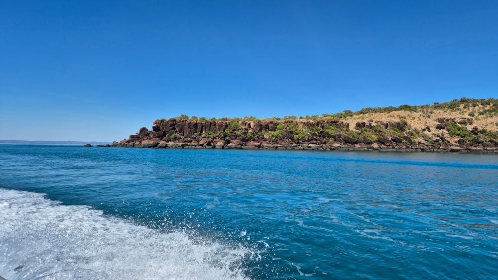 Rocky outcrop with green trees and faded golden grass on a flat and low hill, jutting out of blue ocean. Our boat bow wave in foreground. 