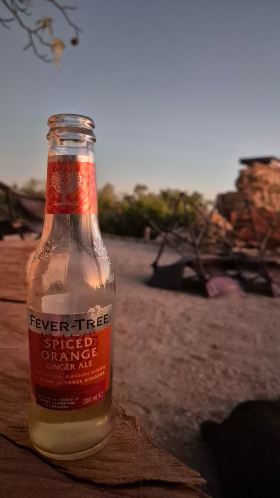 Spiced orange ginger ale cool drink on bench, with large afternoon clear blue sky and some upside down camp chairs in background. 