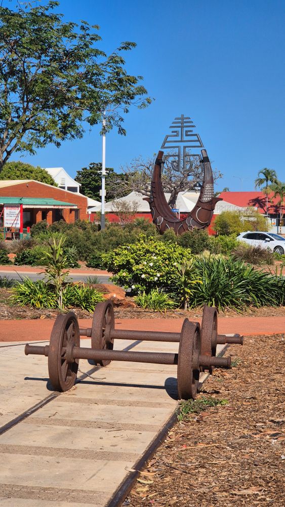Tram base on tracks, in the footpath right in front of greenery with fusion art piece in Broome roundabout. Blue sky and red/terracotta buildings in distance. 