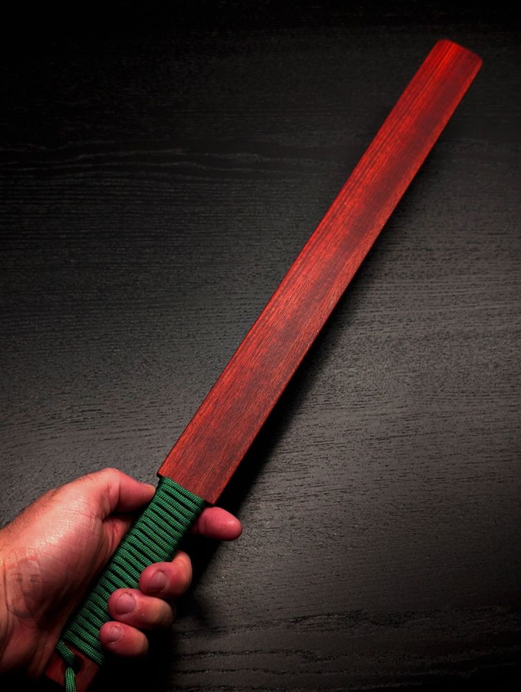 A photo of a wooden spanking paddle made of Bloodwood. The handle is wrapped with green paracord. The grain is a dark red that streaks with lighter shades down the length. It's about 22 inches long and an inch and a half wide with rounded corners at the tip and squared shoulders near the handle. #kink #bdsm #impactplay #impacttoy #woodworking #canadianmaker #smallbusiness #handmade #hardwood #spanking #spanker #paddle #ruler