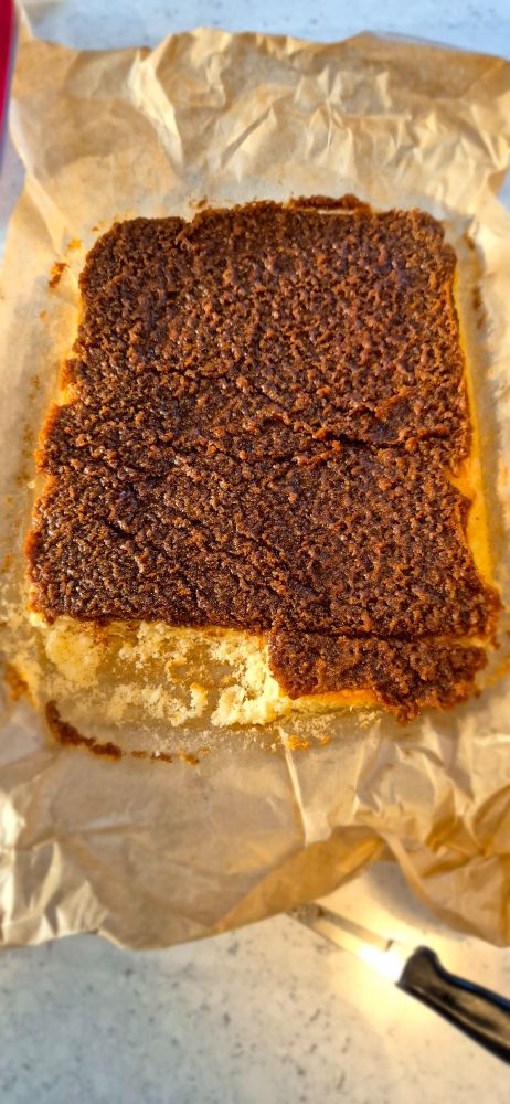 Large rectangular sponge cake. Vanilla sponge with coconut and brown sugar topping.  A slice has been messily cut off at the end. 