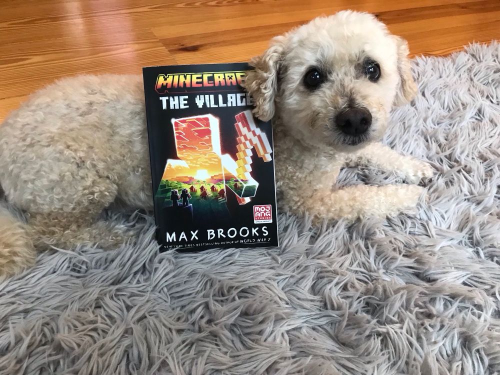 A paperback copy of MINECRAFT: THE VILLAGE posed with a cute little white dog