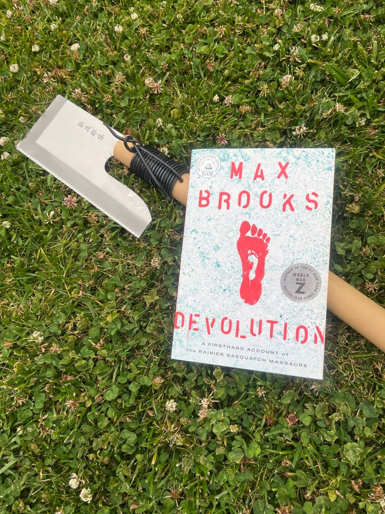 A copy of the book Devolution by Max Brooks lays in the grass on top of a handmade hatchet with a silver blade