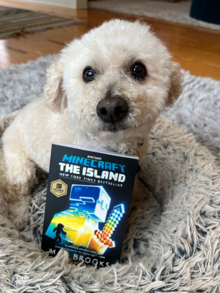 A very cute small white dog named Milo poses with a copy of the book MINECRAFT: THE ISLAND by Max Brooks 