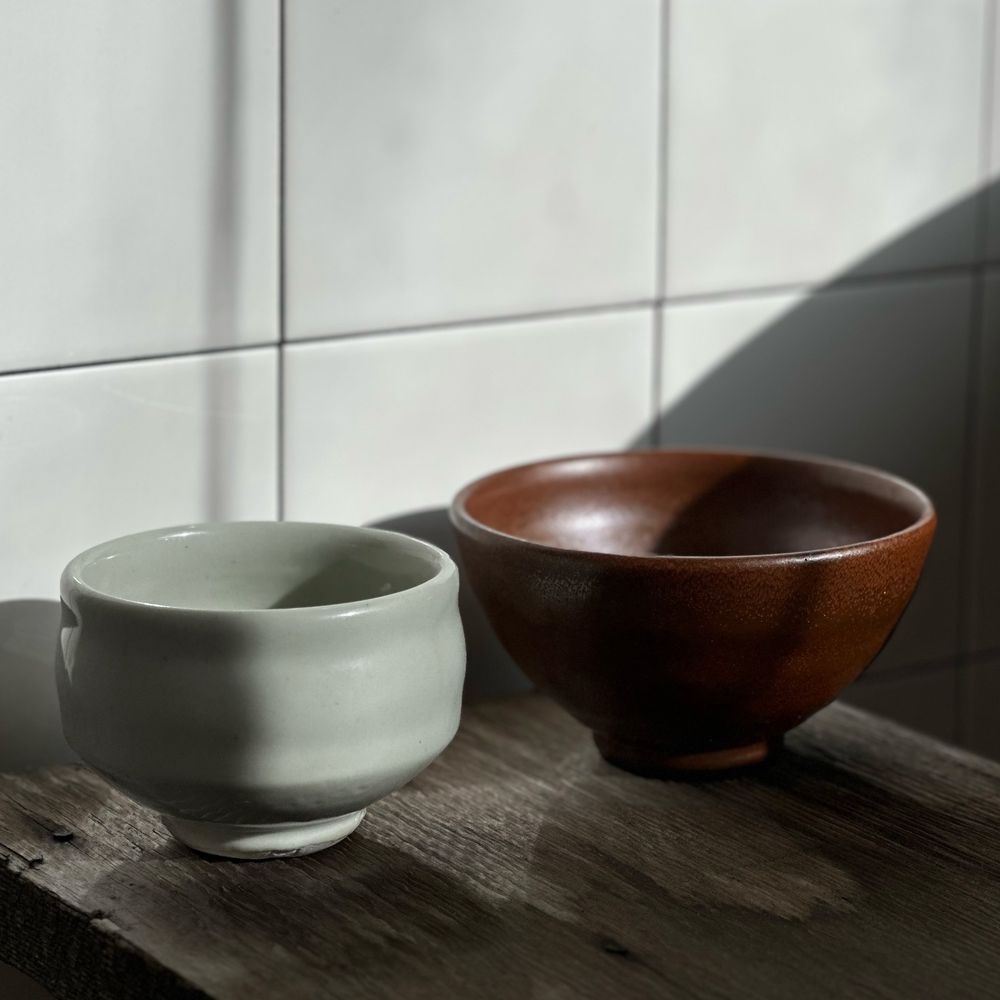 Two bowls in the sunshine and shadows with tiles in the background. One is a pale green with vertical sides the other is a rusty red with a more rounded shape. They are siting on an aged piece of wood.