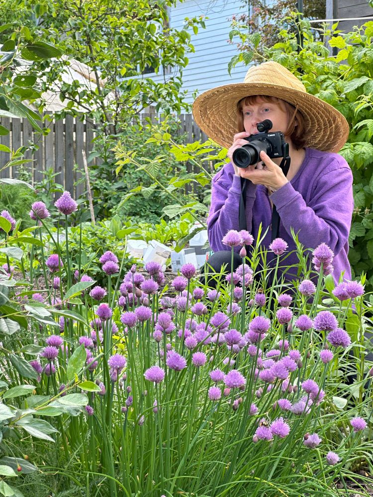 Woman with a wide brimmed hat and red hair wearing a purple sweater holds a camera among purple flowers on a sunny day