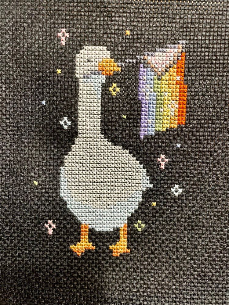 A cross stitched white goose holding a pride flag in his mouth with little sparkles around him on a black background