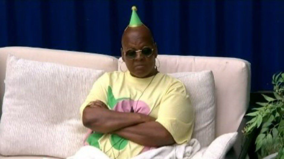 Felicia from bb25 sitting on a white couch with her arms crossed wearing a tiny Green Party hat