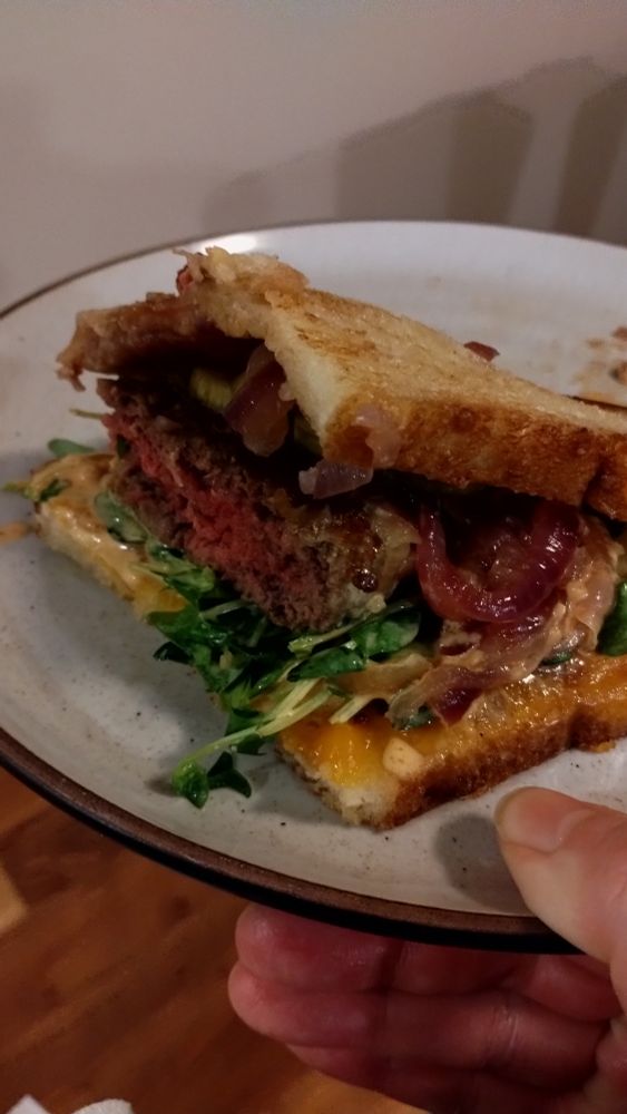 A photo of a patty melt with caramelized onions, pea shoots, pickles, and sriracha mayo alongside a helping of potato chips.