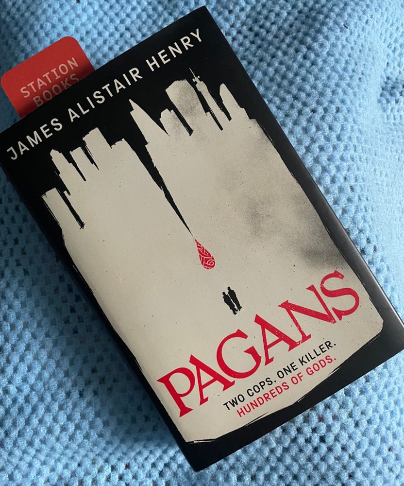 A photograph of a hardback book at a jaunty angle. It is Pagans by James Alistair Henry. Most of the cover is a greyish white, with black around the edges. At the top the shape indicates buildings. The black cuts halfway down the middle of the image in a thin line where a red droplet appears with a traditional old English pattern. This indicates a divide. Below the red droplet are the silhouettes of two people, placed on what would be either side of the divide if it went that far.
The title of the book is written in red.
Along the bottom of the cover, in two lines, are the words: two cops, one killer, hundreds of gods.