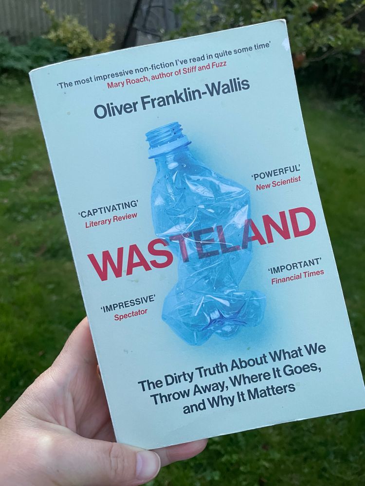 A photograph of a paperback book, in a hand, in a garden. The cover is light blue and contains an image of a squashed, blue-tinged plastic bottle that I assume would’ve contained water. It includes the words: Oliver Franklin-Wallis, Wasteland, and a few quotes of praise for the book.