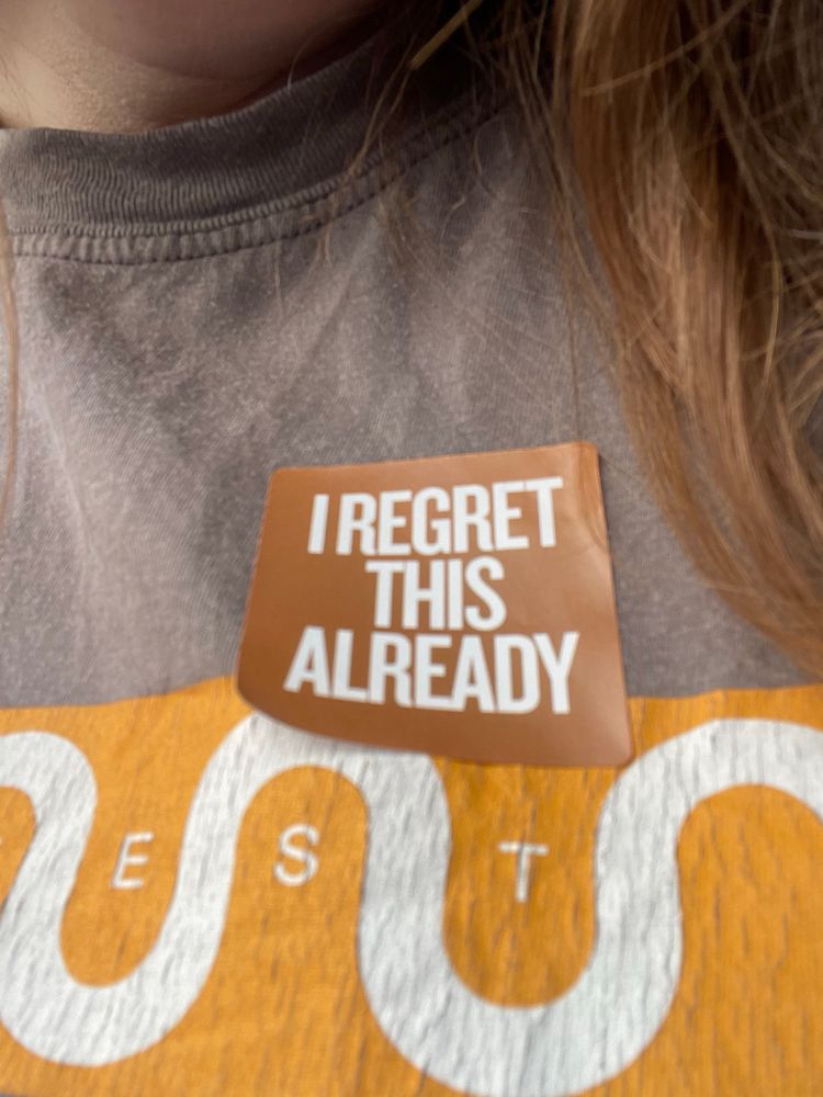 A close up photo of a square sticker containing the words: I regret this already. The sticker is stuck on my T-shirt.