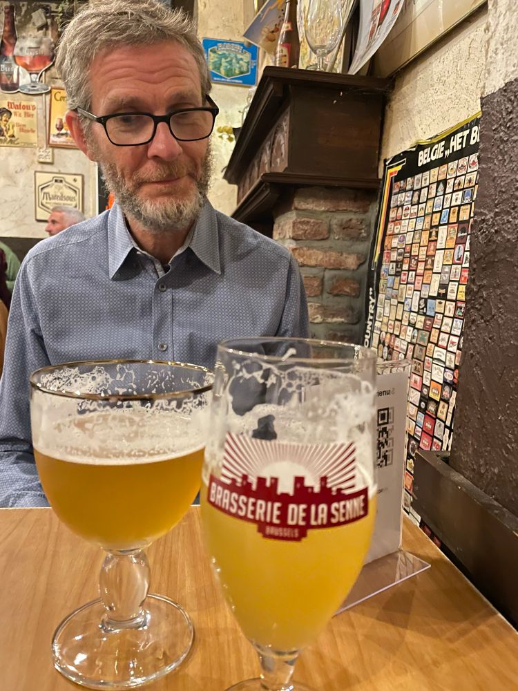A photo of a man in a blue shirt looking quizzically at two glasses of beer. The glasses are both differently shaped, rounded glasses.