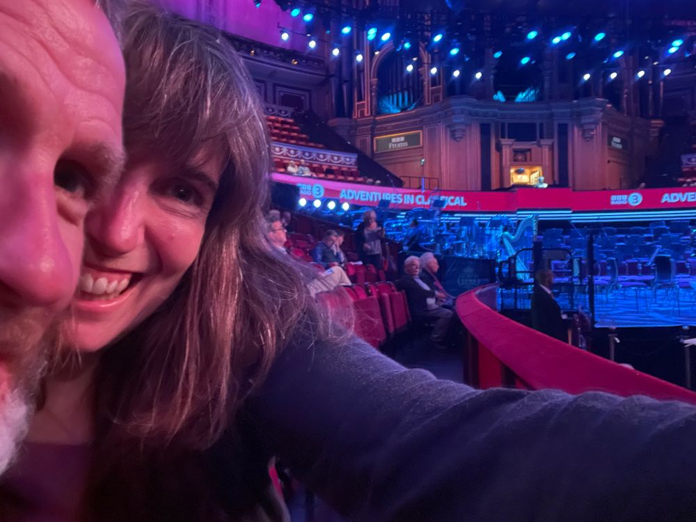 A bad selfie. From left we can just about see a man’s face, behind him is half a woman’s face. Both are smiling. The woman’s arm reaches in front of the rest of the view, as she holds the phone to take the selfie. The rest of the photo shows a railing, and in the distance at the same level, the stage inside the Royal Albert Hall.