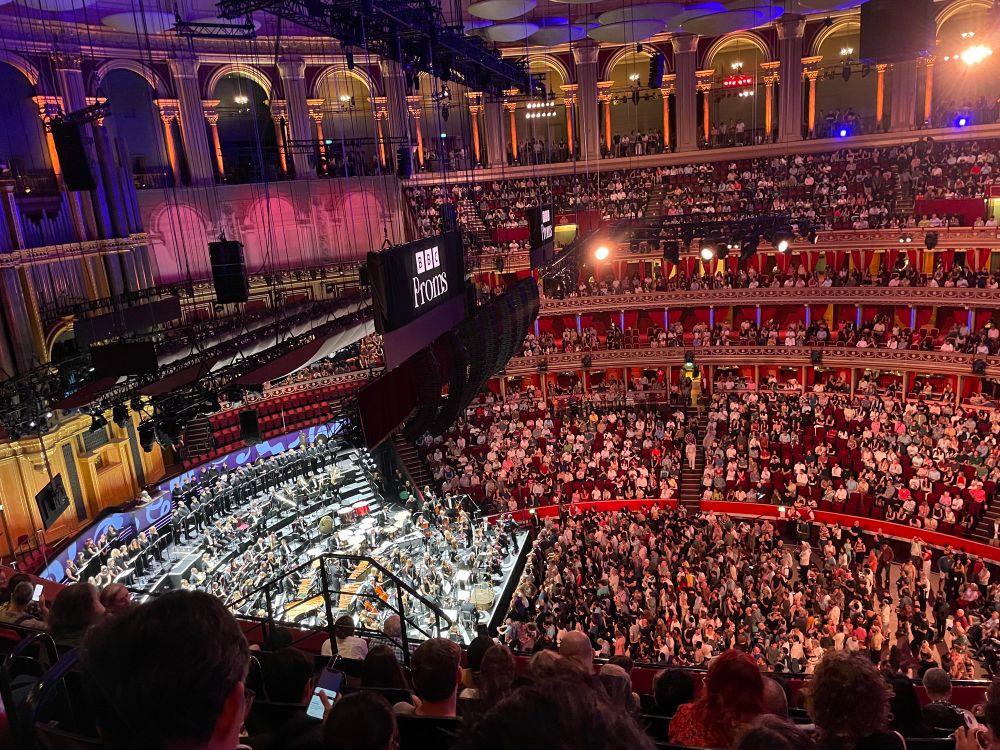 A photograph looking down from the top row of the Rausing Circle in the Royal Albert Hall. Hundreds of people are in the audience, which is illuminated red. To the left we can see an area of people illuminated white, this is the orchestra. If you zoom in you can just about see that the percussion section has been moved to the middle of the orchestra. But everything is very far away so it’s hard to tell.