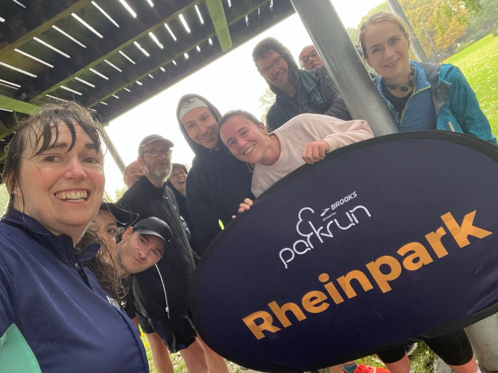 A selfie of a group of rain-soaked but smiling people holding the sign for Rheinpark parkrun.