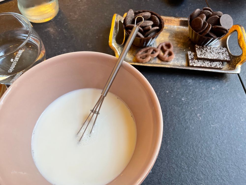 A photograph looks more king down into a large, almost bowl-like mug containing warm milk and a miniature whisk. To one side is a small tray containing two dark chocolate pots of large, dark chocolate buttons to pour into the milk.
