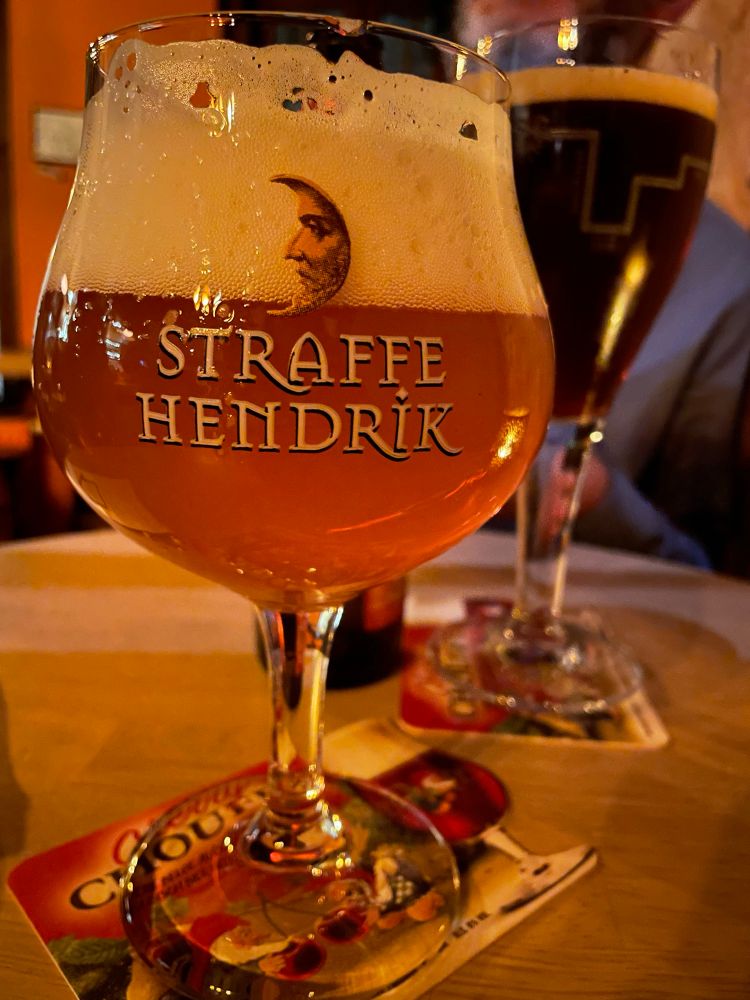 A round glass of beer. It bears the words Straffe Hendrick and contains a warm, golden brown beer. In the background is a tulip glass containing a dark brown beer.