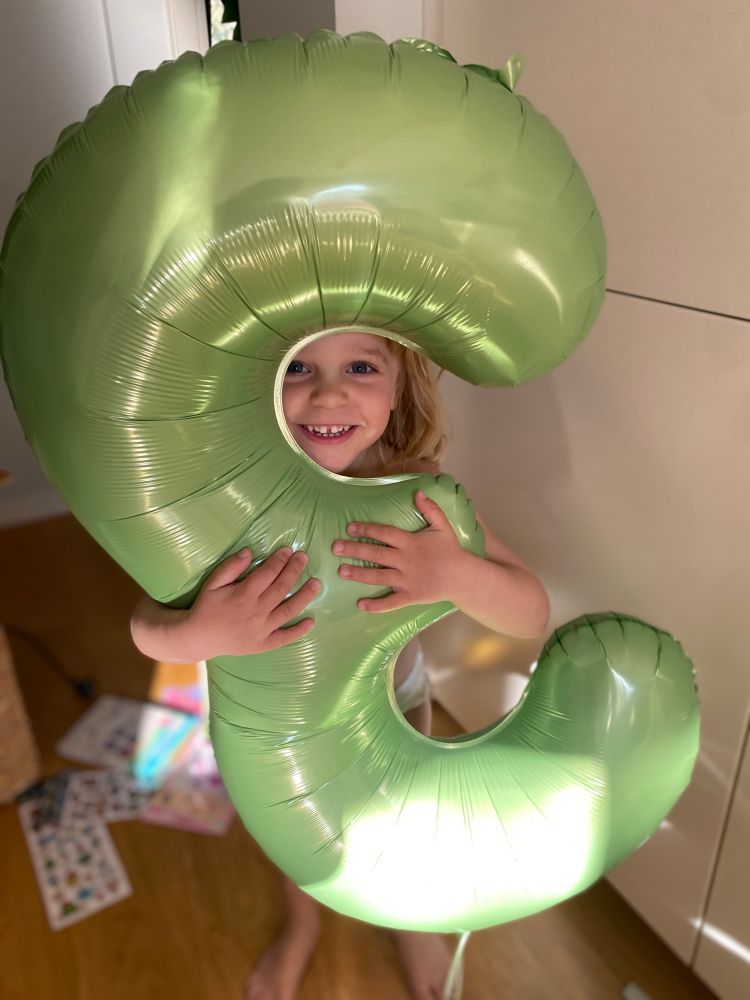 En treåring står tittar fram bakom en stor grön heliumballong i form av en trea. Hon kramar om ballongen och ser glatt in i kameran. 