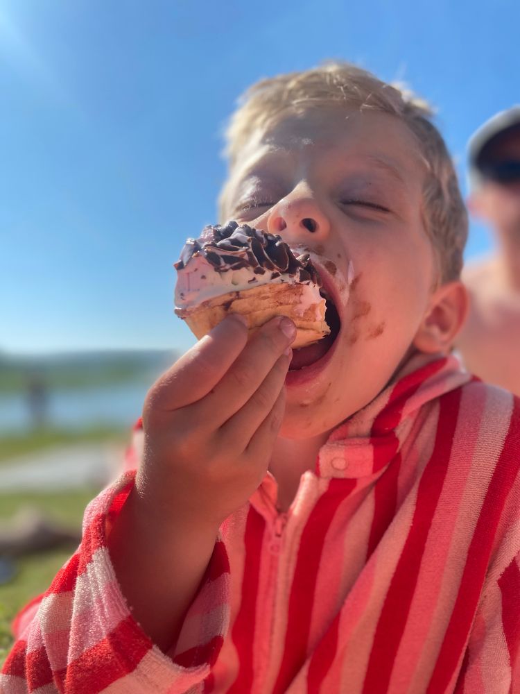 Närbild på ett blont femårigt barn som äter glassbåt med en stor tugga. Det är blå himmel, soligt och barnet har badrock på sig. 
