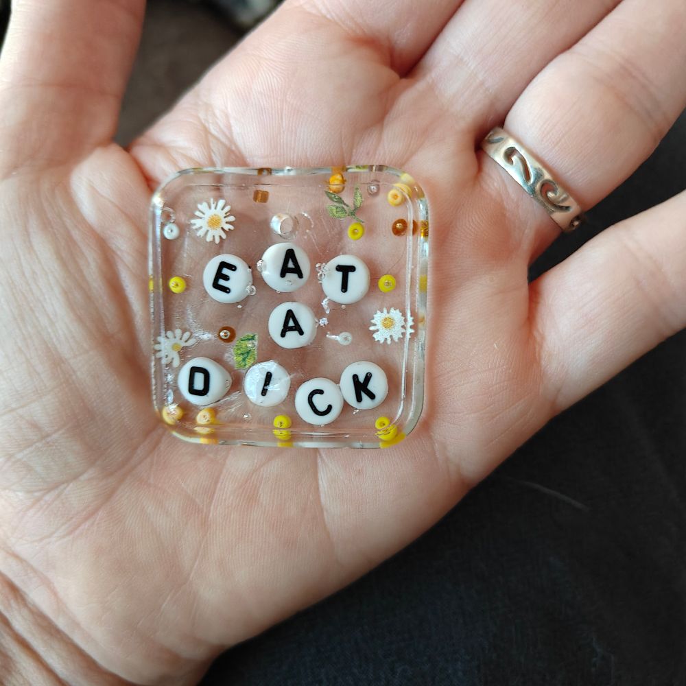Little resin pendant within bedded flowers and beads that says eat a dick. The letters kind of floated up and are all wonky and there's some bubbles.