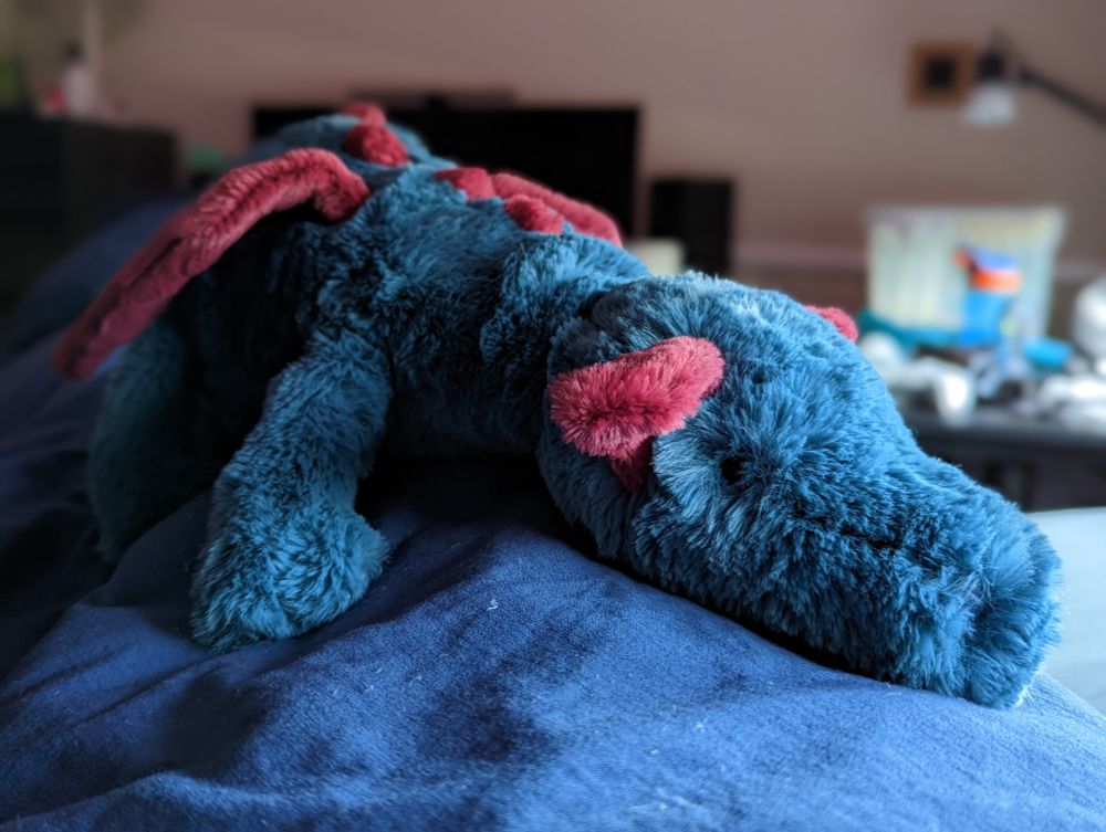 A fuzzy dragon plushie laying on a blanket, looking at the viewer.