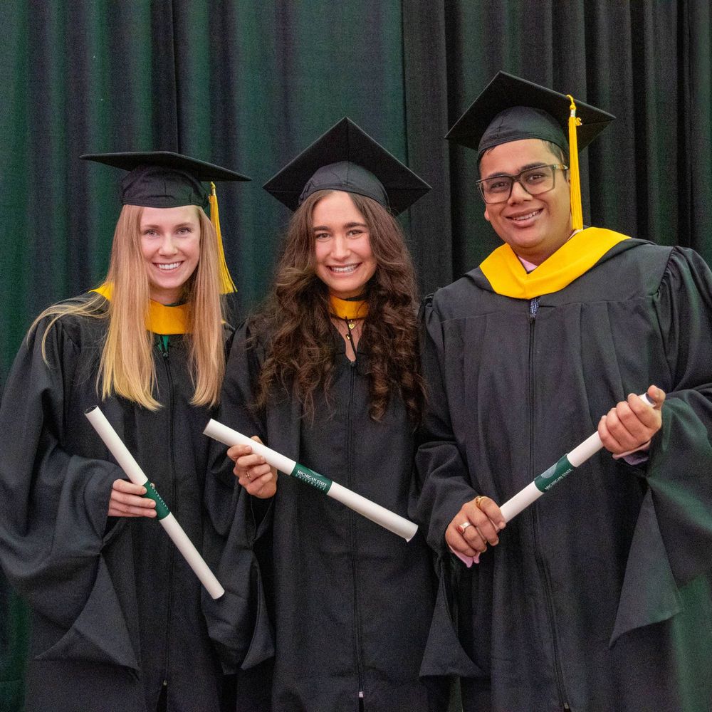 Three masters' students stand in their caps and gowns with their diplomas in their hands. 
