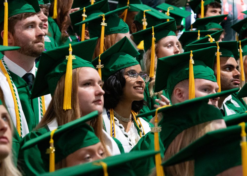 A group of MGI undergrads in green caps and gowns looks up. One girl in particular has a smile on her face and is looking up. Her face is featured in the picture. 
