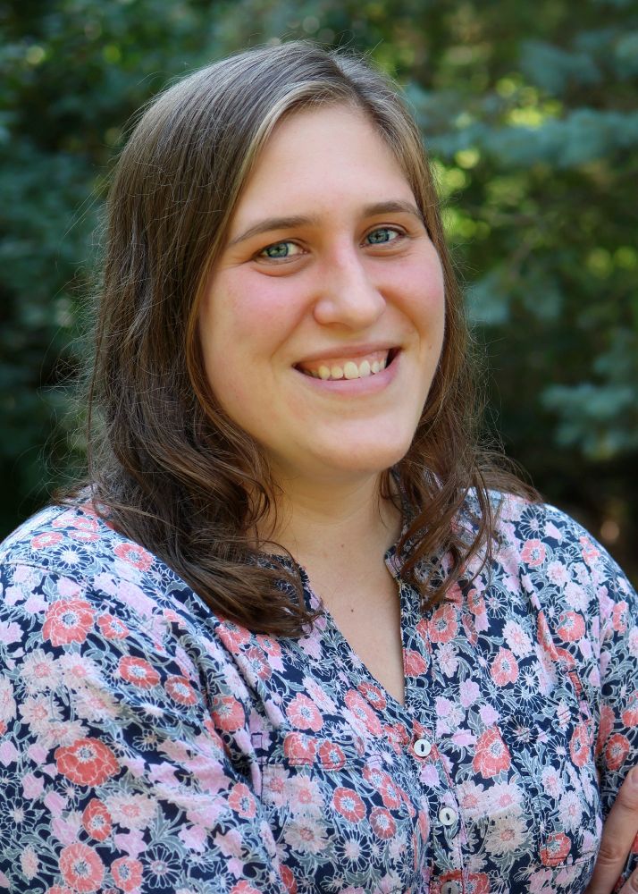 Elizabeth Heath-Heckman stands outdoors and smiles at the camera. She is wearing a blue, coral, pink, and white floral shirt. 
