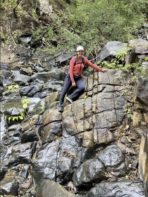 Sarah Gonzalez sits on a rock wall. The wall looks wet, and it extends up behind her as well. There are plants growing among the rocks.  She's wearing athletic clothing, a backpack, and a baseball cap.