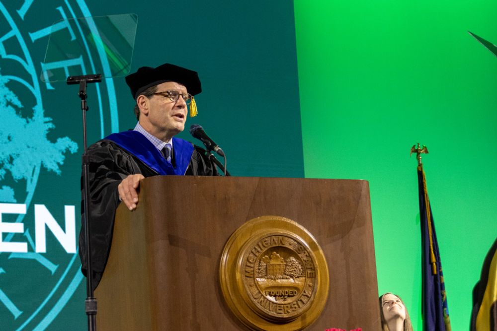 MGI Chair Vic DiRita stands behind a lecturn with the MSU seal on it. He is wearing regalia. 
