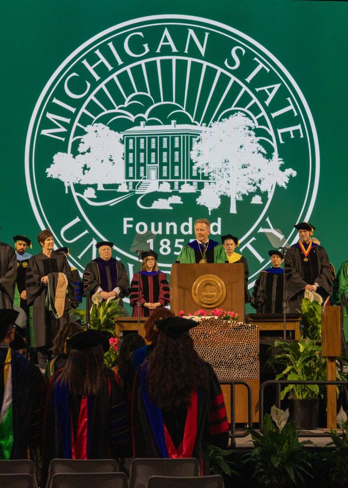 MSU President Guskiewicz gives an address at the Masters graduation ceremony. He's standing behind a lecturn with a large MSU seal displayed behind him. He and everyone on the stage are wearing regalia. 
