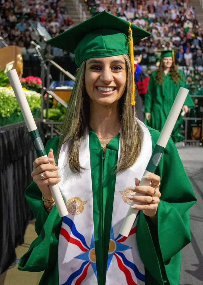 A new graduate in green cap and gown with a multicolored stoll holds two diplomas and has a huge smile on her face. 
